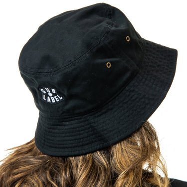 Black bucket hat with 'Sub Label' logo worn by a girl with wavy hair on a white background