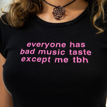 Black t-shirt with pink text 'everyone has bad music taste except me tbh' worn by a person.