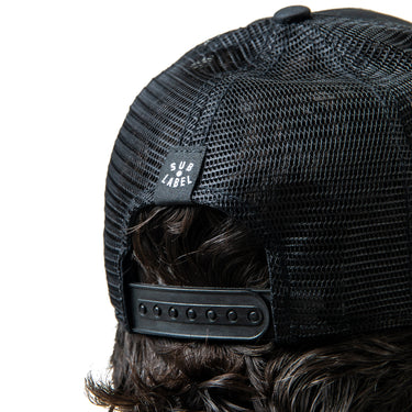 Black mesh trucker cap with a brand label worn by a person with wavy hair.