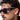 Close-up of a person wearing black sunglasses with a brand logo, against a neutral background.