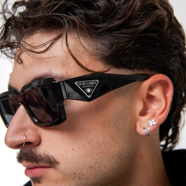 Close-up of a person wearing black sunglasses with a brand logo, against a neutral background.